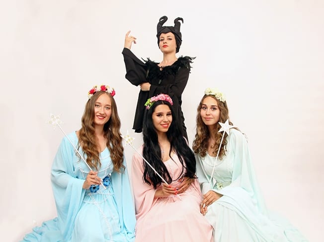 Trei interprete în rochii pastel așezate și o a patra interpretă în negru/coarne în picioare.